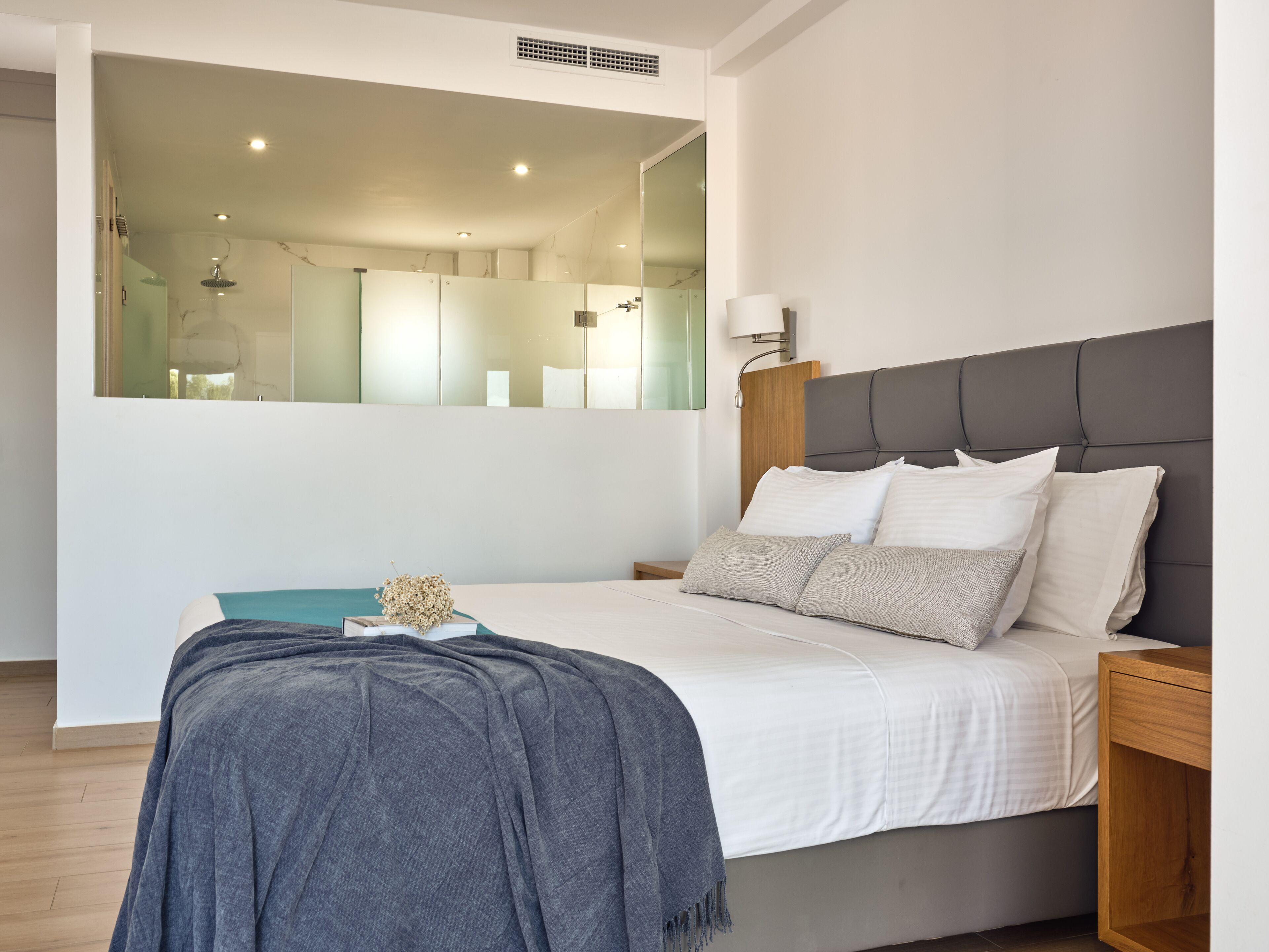 A modern bedroom with a large bed, a nightstand, a mirror, and a wall-mounted light.