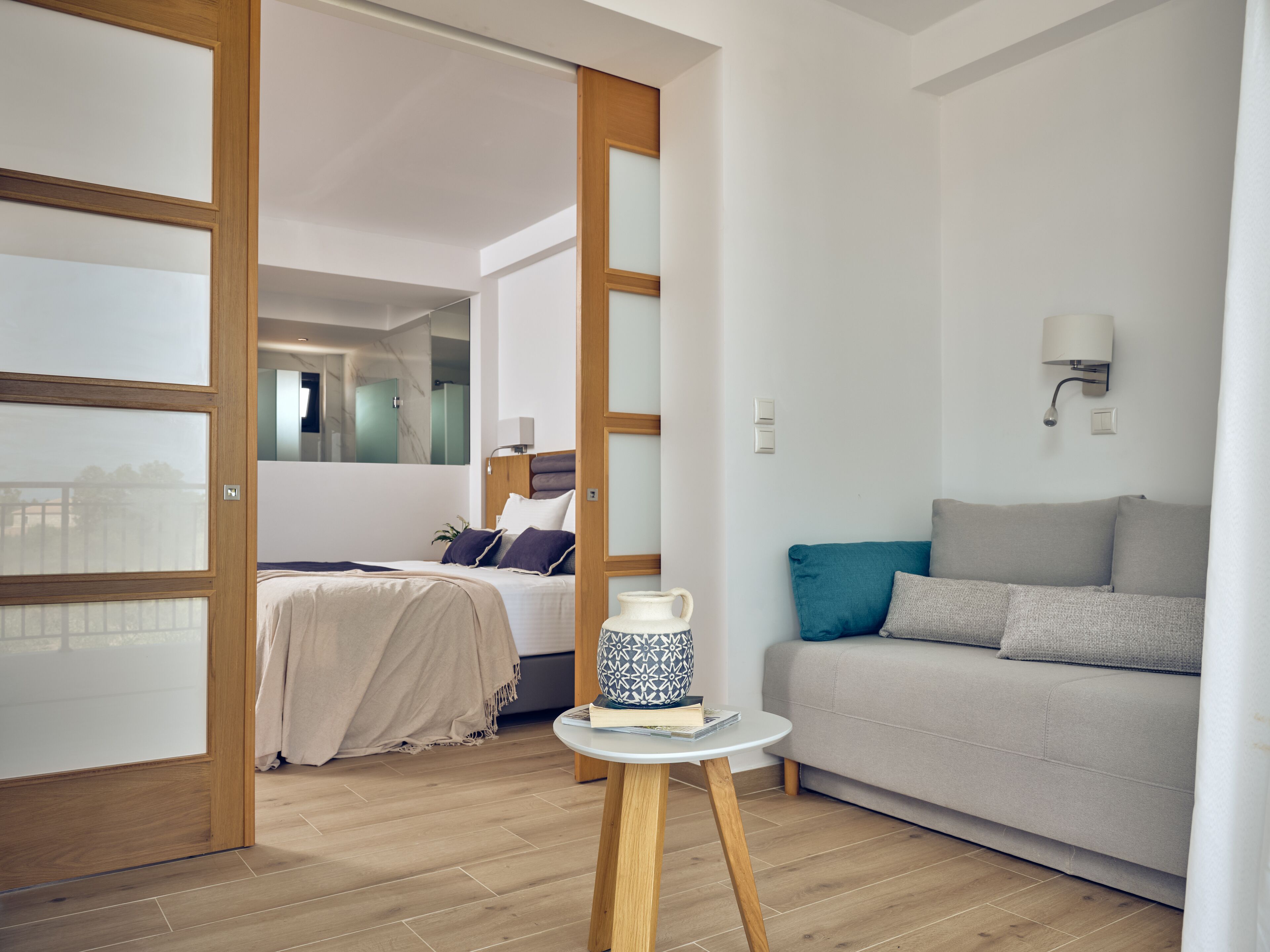 A modern hotel room with a bed, a sofa, a small table with a decorative item, and a mirror.