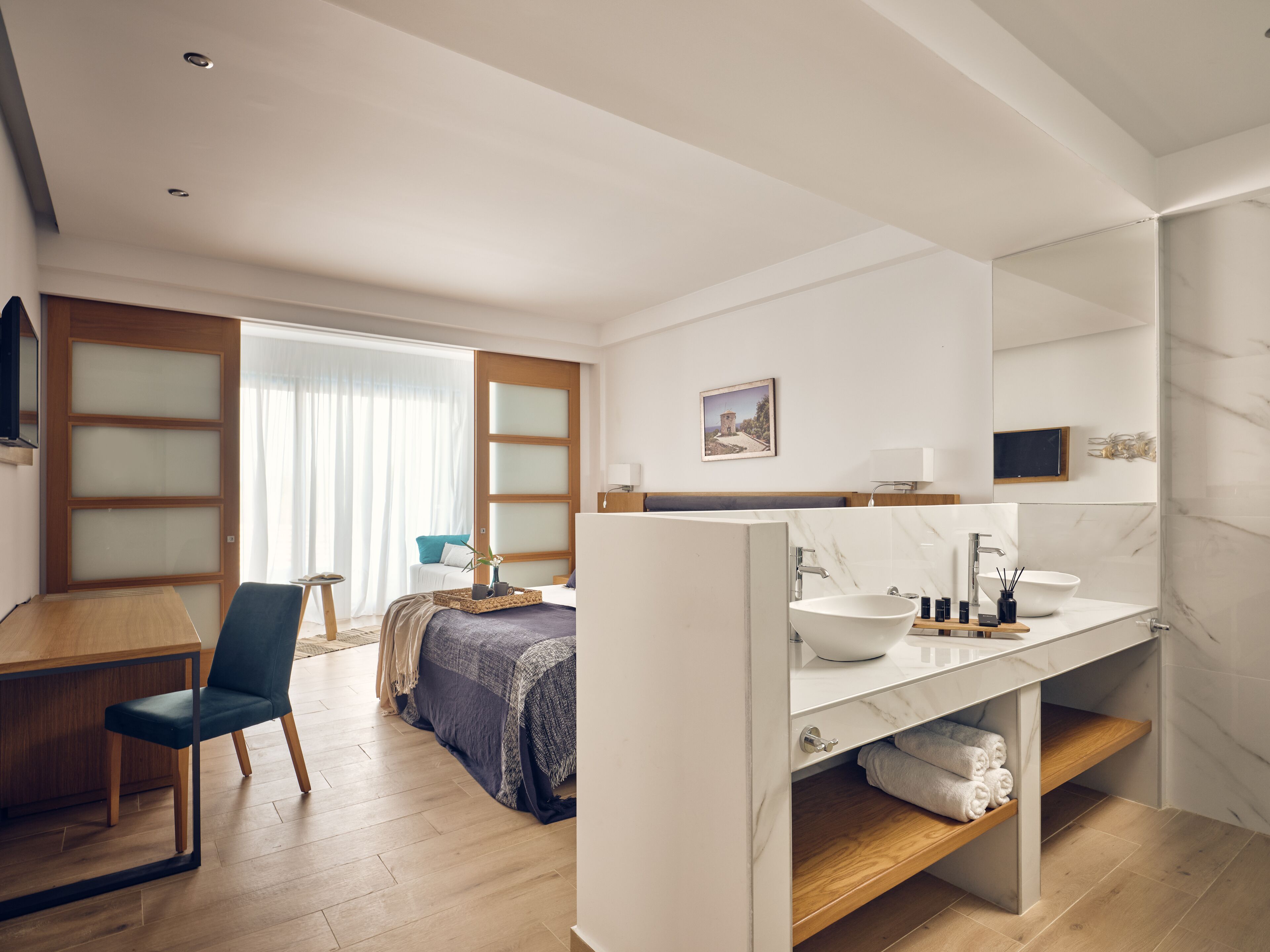 A modern hotel room with a wooden desk, a comfortable chair, a bed with a blue blanket, a large window with curtains, a bathroom with a marble sink, and a small bedside table with a lamp.