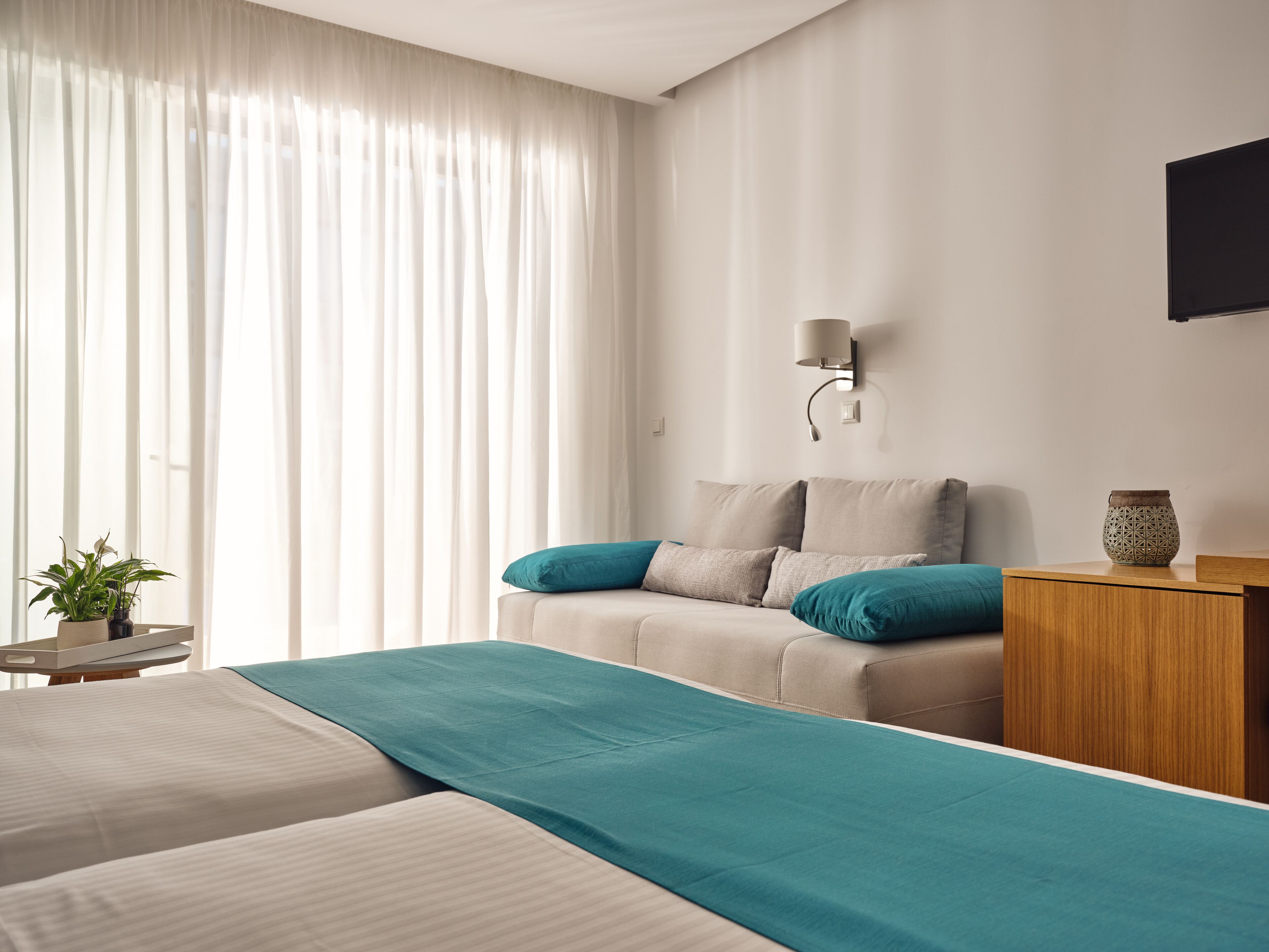 A modern hotel room with a large bed, a bedside table, a lamp, and a view of the outdoors through sheer curtains.