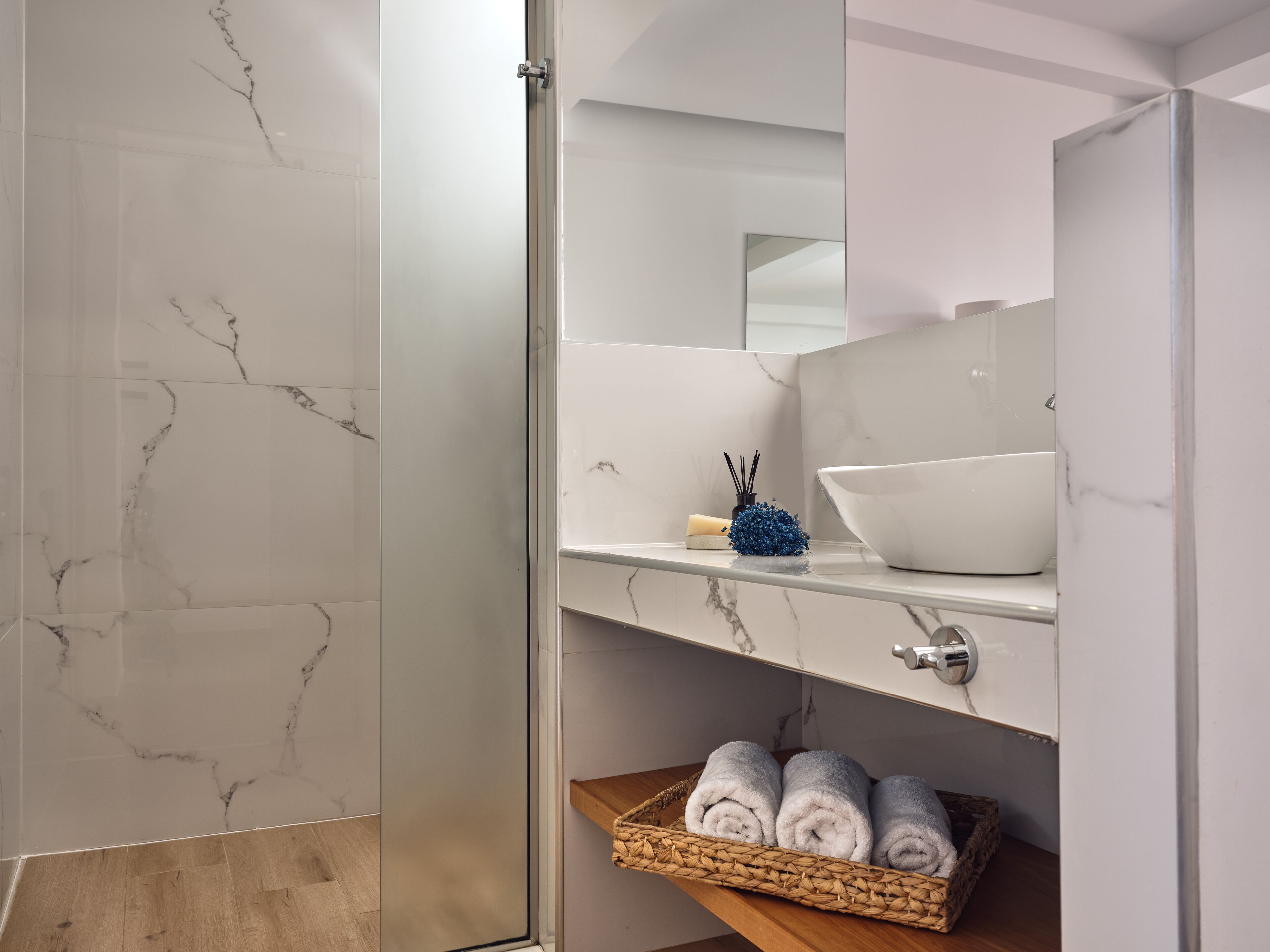 Un baño con piso de mármol, un lavamanos blanco, un espejo y una repisa de madera con toallas.