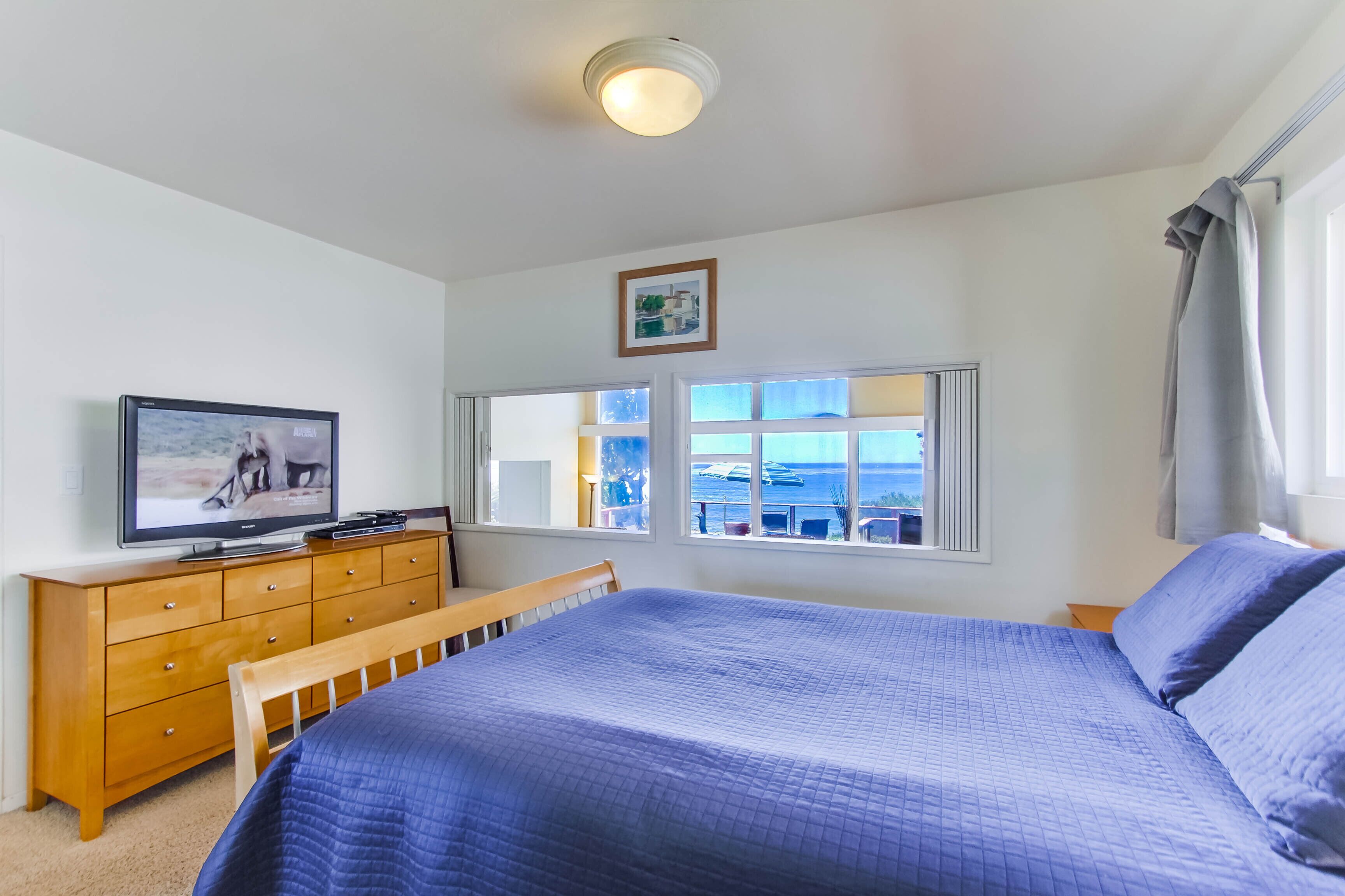 A bedroom with a bed, a wooden dresser, a television on the dresser, a window with a view, and a ceiling light.