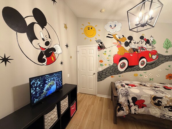 A room with a wall mural of Mickey Mouse and Minnie Mouse, a television, a bed with a Mickey Mouse-themed duvet, and a door.