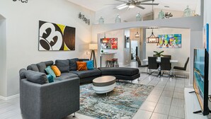 A modern living room with a sectional sofa, a coffee table, and a dining area with a chandelier. - Unique Disney Villa 5BR, Heated pool, Hand-Painted Murals, 3 Miles to Disney (Kissimmee)