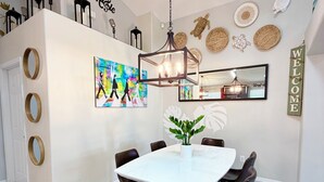 A modern dining area with a white table, black chairs, a hanging light fixture, a 'Welcome' sign, and a colorful abstract painting. - Unique Disney Villa 5BR, Heated pool, Hand-Painted Murals, 3 Miles to Disney (Kissimmee)