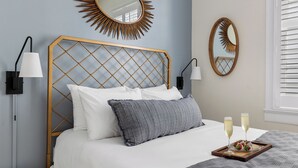 A neatly made bed with white and gray pillows, two bedside lamps, a sunburst mirror, and a tray with champagne glasses and strawberries.