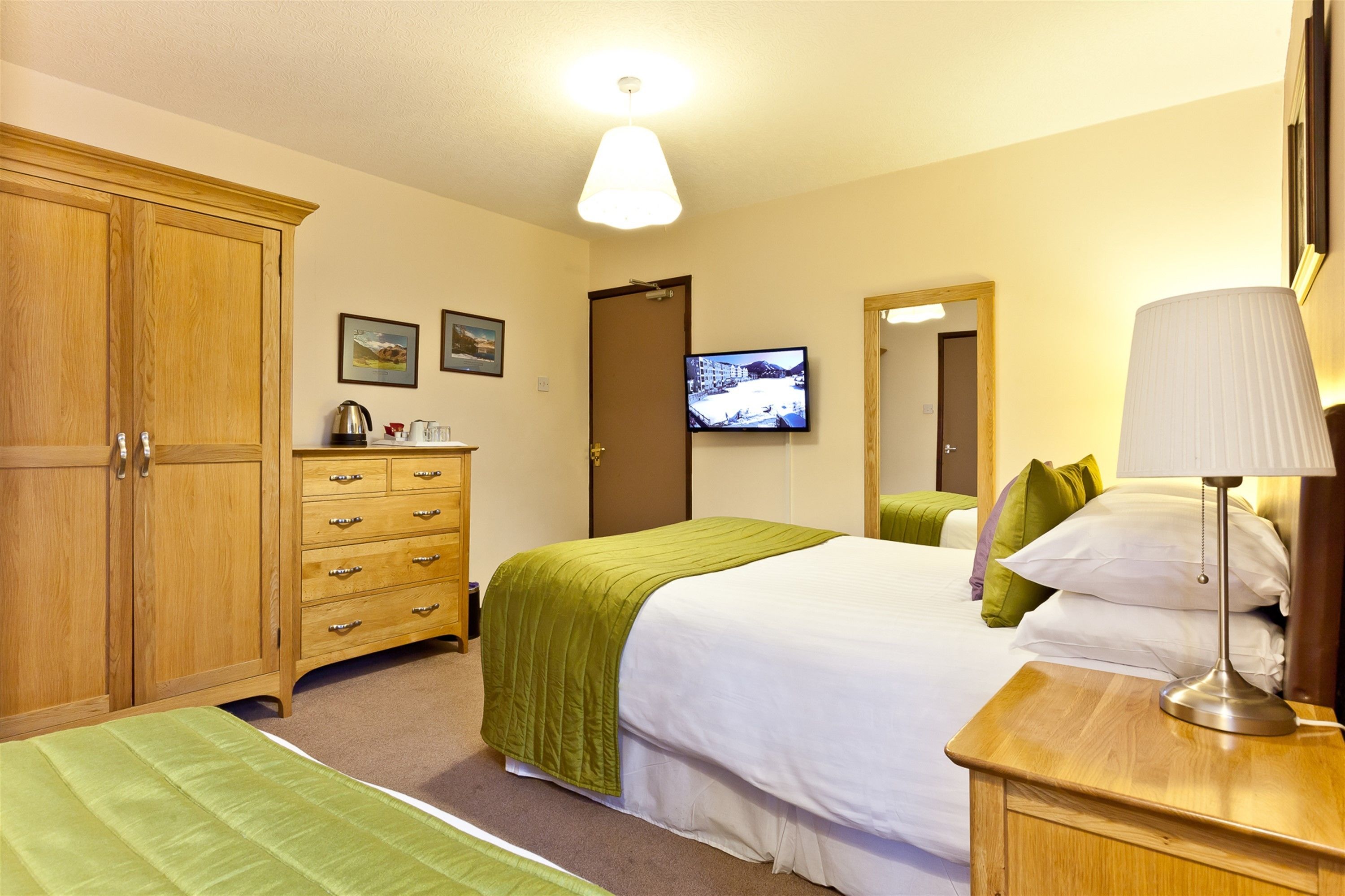 A hotel room with a large wooden wardrobe, a bed with green bedding, a bedside table with a lamp, and a television on the wall.