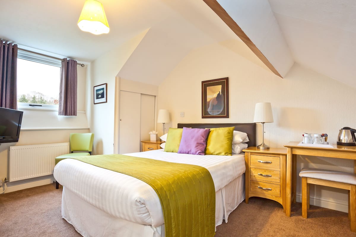A bedroom with a bed, bedside tables, a desk, a chair, a television, and a window with curtains.