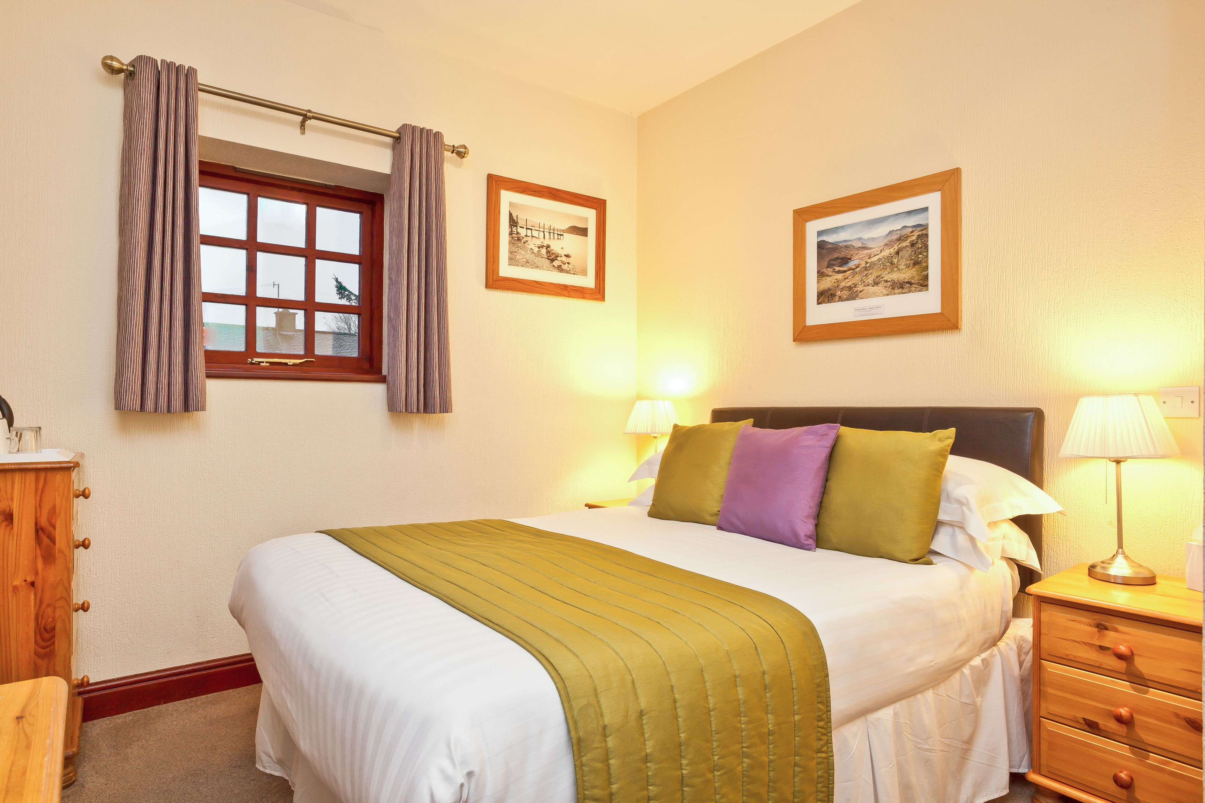Camera d'albergo ordinata con un letto, abat-jour, un comodino in legno e due quadri appesi al muro.