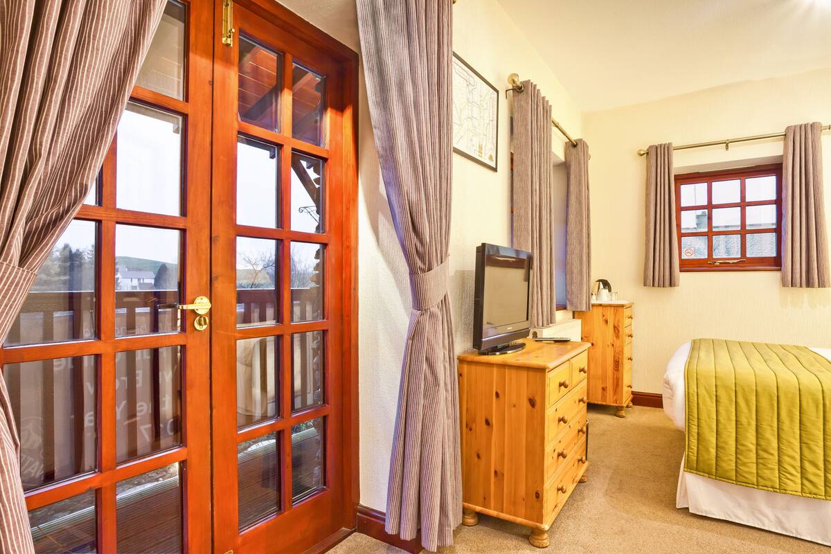 A bedroom with a wooden dresser, a television on top, a bed with a green blanket, and a window with curtains.