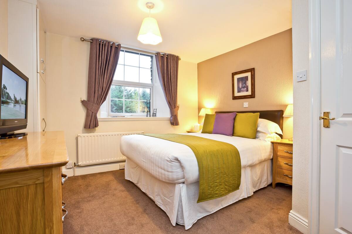 A bedroom with a bed, bedside table, lamp, window with curtains, radiator, and a television on a stand.