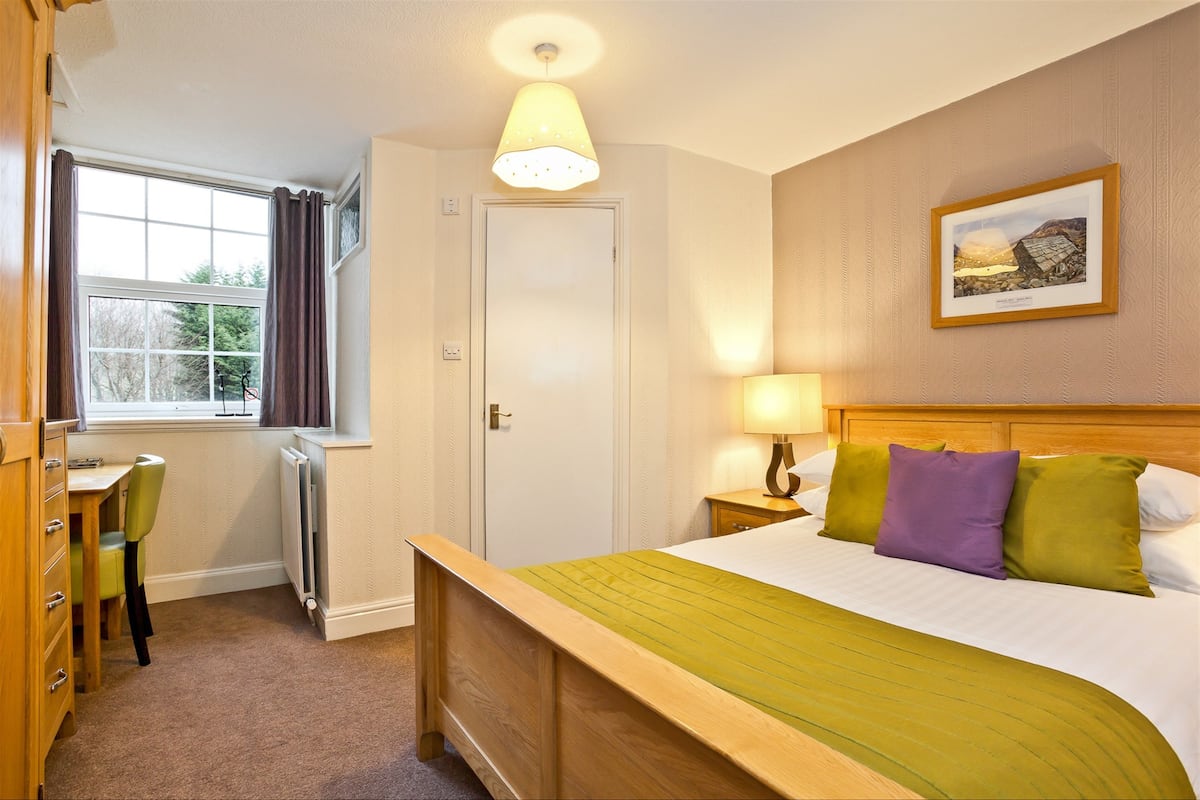 A bedroom with a wooden bed, a bedside table with a lamp, a window with curtains, and a radiator.