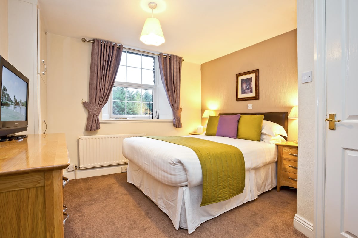A bedroom with a bed, bedside table, lamp, window with curtains, and a television on a stand.