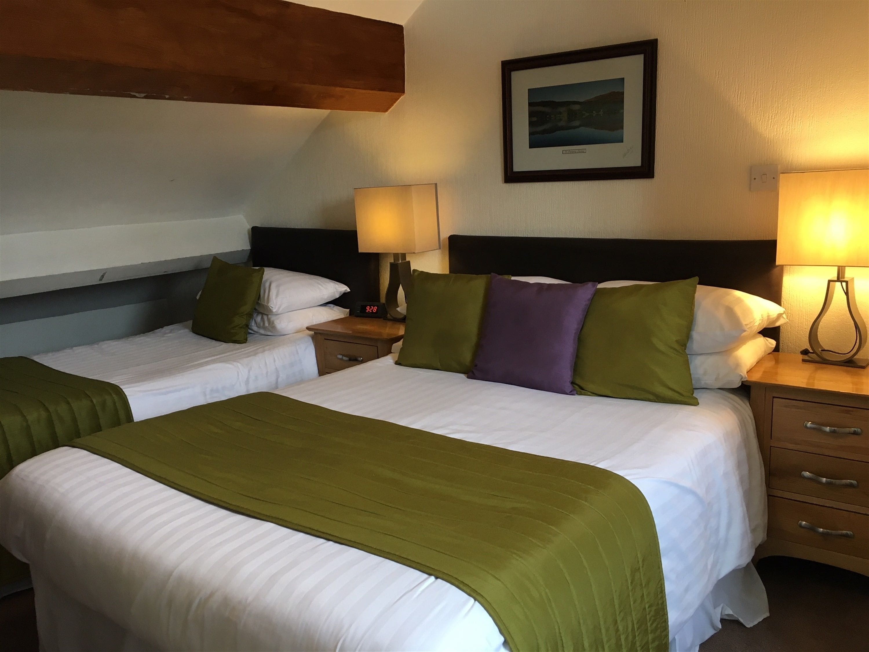 Une chambre d’hôtel avec deux lits, chacun recouvert d’une couverture verte et doté de draps blancs et d’une tête de lit. On y trouve des tables de chevet avec des lampes, une horloge et un tiroir. Un tableau encadré est accroché au mur, au-dessus des lits.