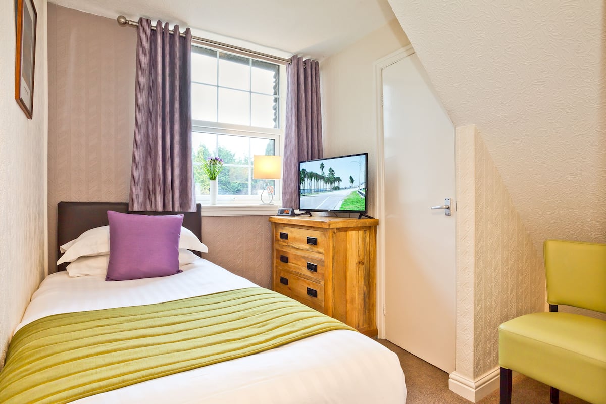 A bedroom with a bed, a wooden dresser, a television, a window with curtains, and a chair.
