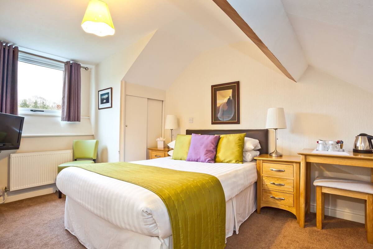 A bedroom with a bed, bedside tables, a desk, a chair, a television, and a window with curtains.