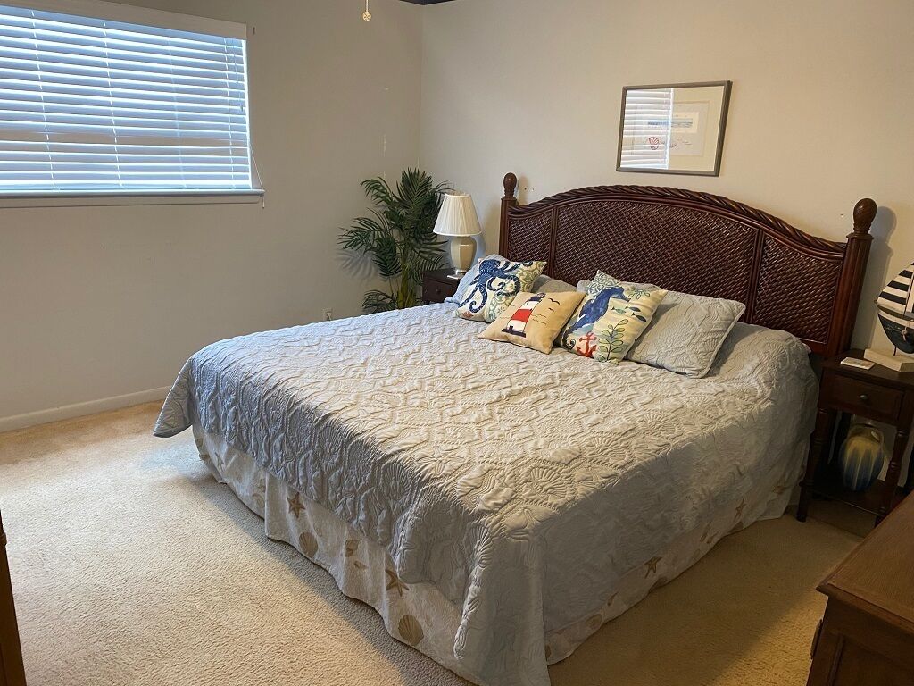 A bedroom with a large bed, a nightstand, a lamp, a framed certificate on the wall, and a window with blinds.