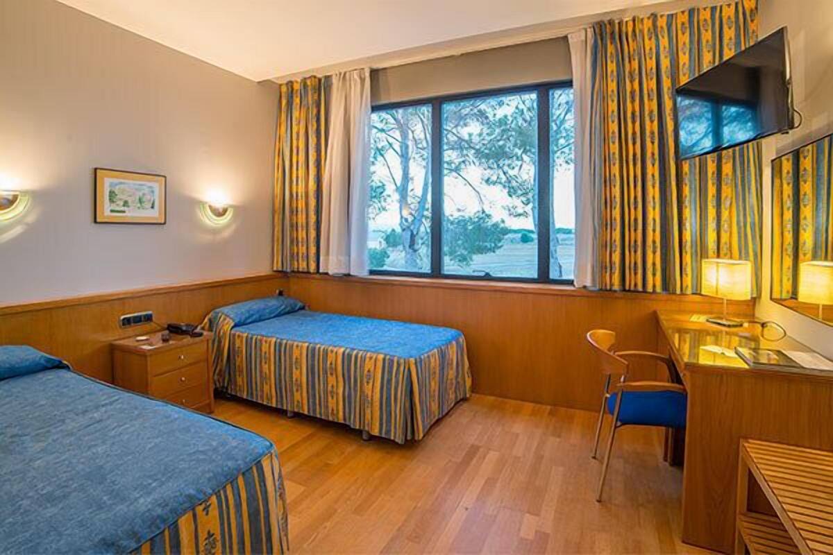 A hotel room with two beds, a desk, a chair, a TV, and a window with curtains.