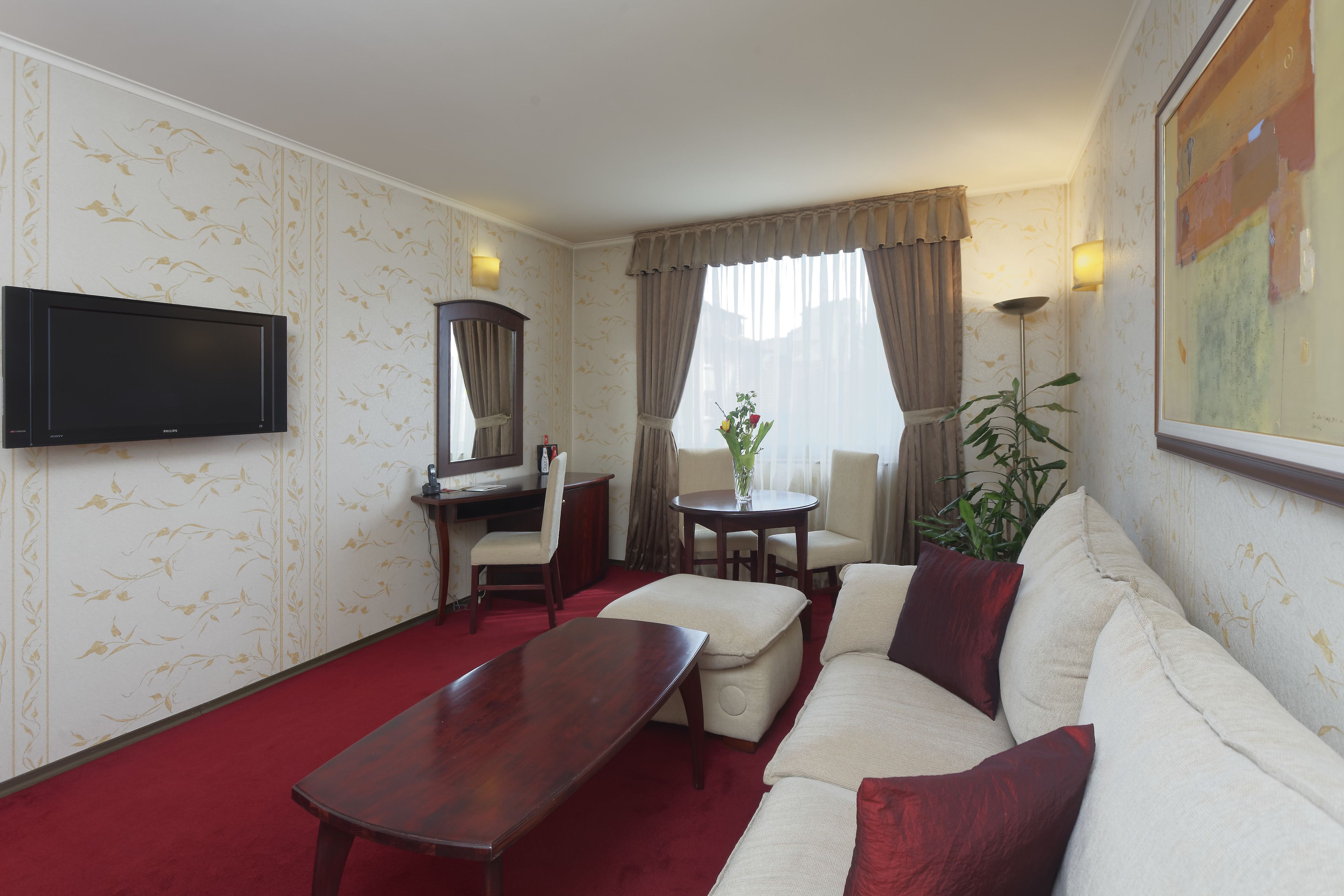 A hotel room with a red carpet, a sofa, a table, a desk with a chair, a TV mounted on the wall, and a window with curtains.