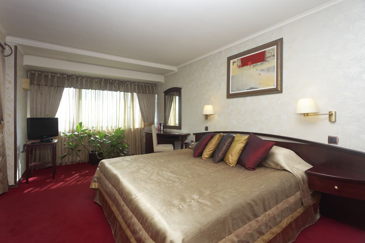 Quarto de hotel com uma cama grande, uma escrivaninha com televisão, duas luminárias de cabeceira e um quadro na parede.
