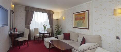 Une chambre d’hôtel avec un tapis rouge, un canapé, une table basse, un bureau et une chaise. Il y a un tableau au mur et une plante dans un coin.