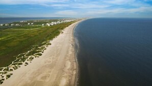 Eine Vogelperspektive auf eine Küstenregion mit einem Sandstrand, grünen Feldern und einer Reihe weißer Gebäude entlang des Ufers.