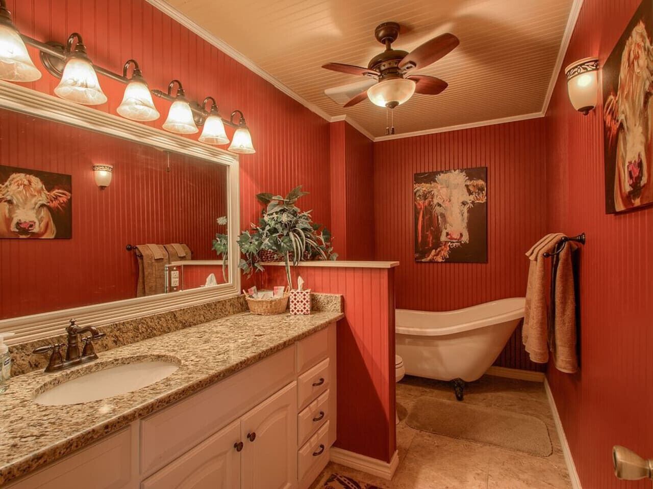 A bathroom with a red wall, a white bathtub, a granite countertop sink, a ceiling fan, and a mirror with lights.