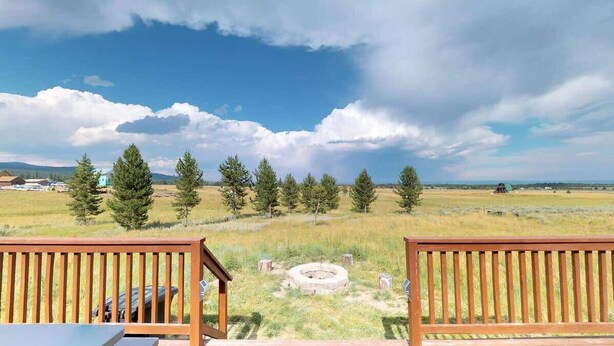 A wooden deck with a view of a grassy field, trees, and a distant building. - 32 MI to YNP | Pet Friendly | Sleeps 2 (Island Park)