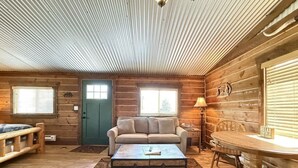 A cozy cabin interior with a wooden floor, a sofa, a coffee table, a dining table with chairs, and a lamp. - 32 MI to YNP | Pet Friendly | Sleeps 2 (Island Park)