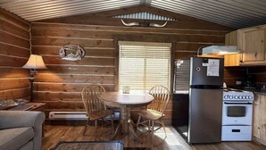 A cozy cabin interior with a wooden dining table, chairs, a refrigerator, and a stove. - 32 MI to YNP | Pet Friendly | Sleeps 2 (Island Park)