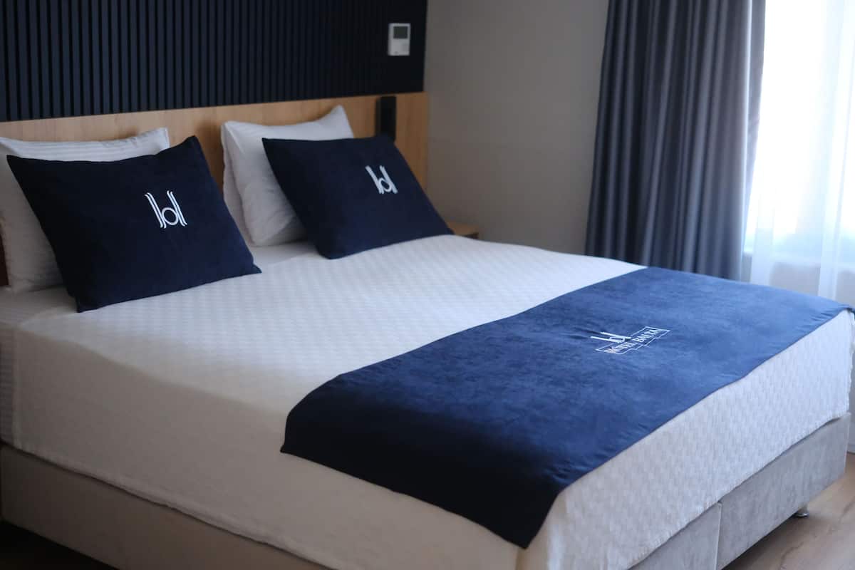 Una cama bien tendida con una colcha blanca y azul marino, dos almohadas azul marino con un logotipo, y un cabecero de madera.