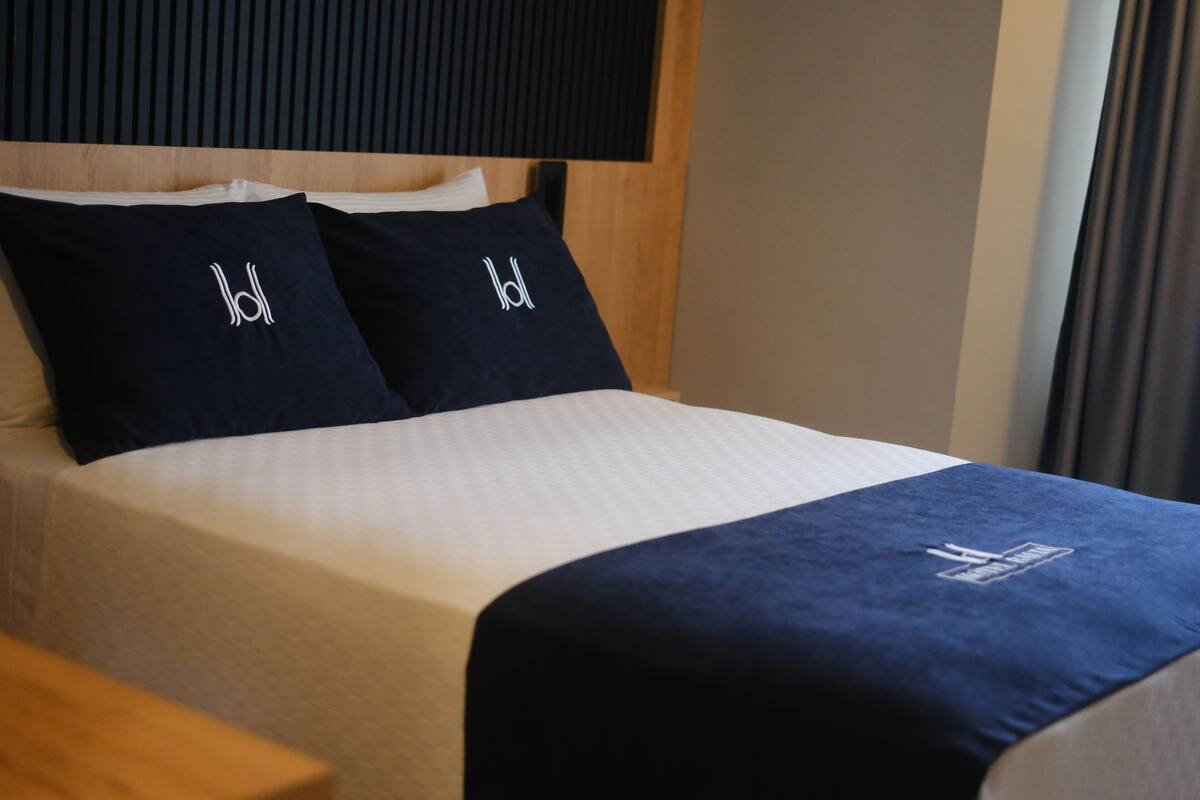 Una cama bien hecha con dos almohadas que llevan un logotipo, un cabecero de madera y una manta azul oscuro.