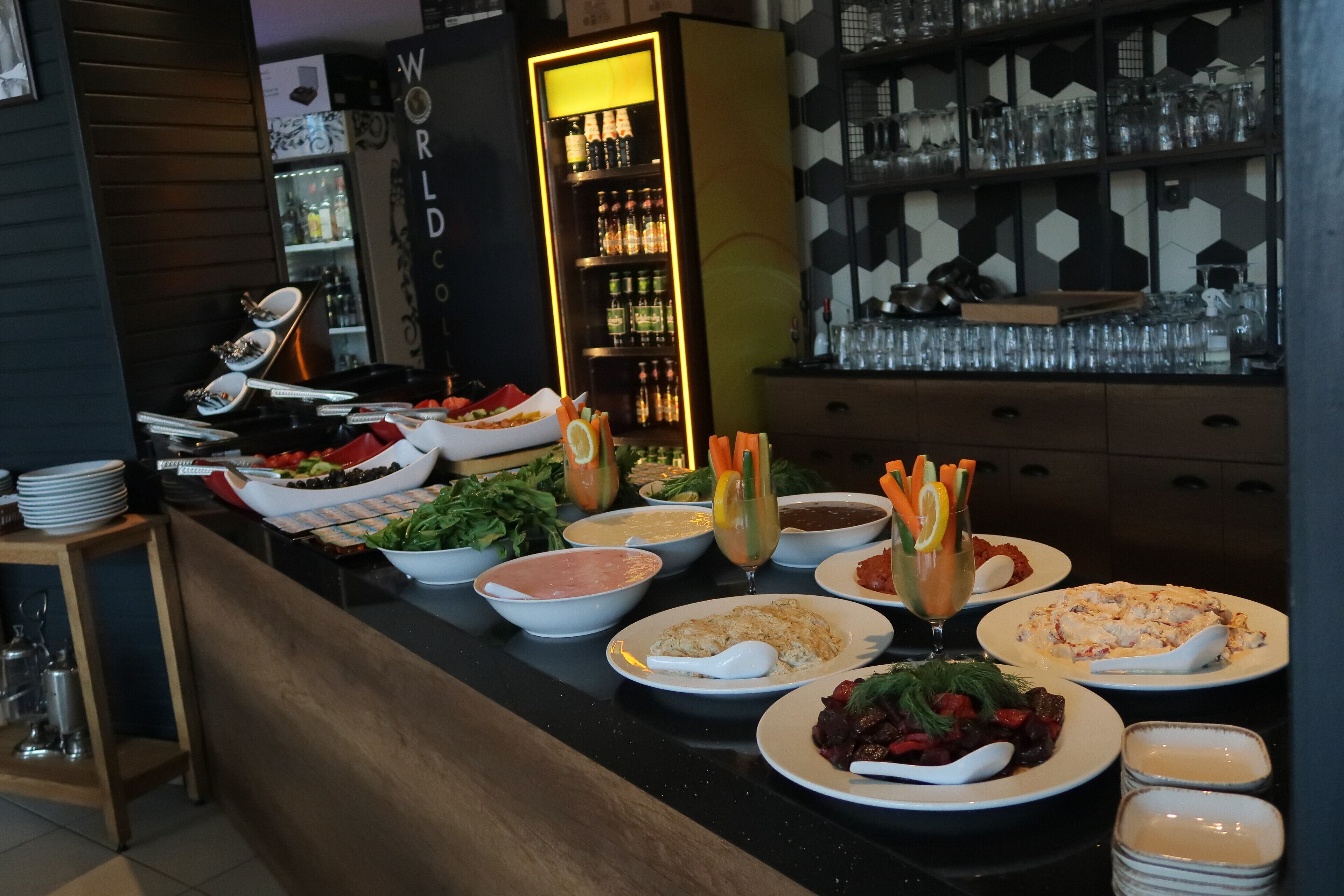 Un bufé con diversos platos y bebidas, un refrigerador con botellas, y una pared con diseño.