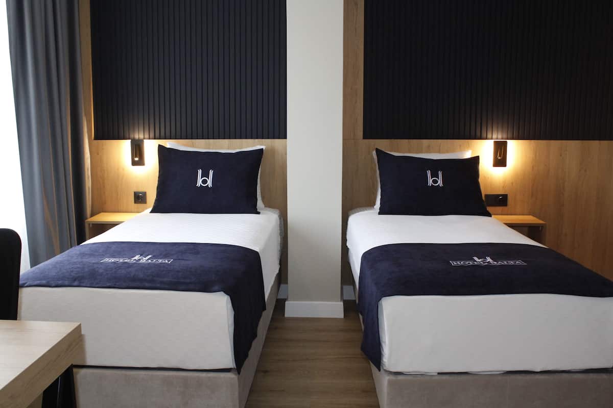 Habitación de hotel con dos camas, cada una con cabecera y almohada azul oscuro, y una colcha blanca. Hay una mesita de noche de madera con una lámpara entre las camas.