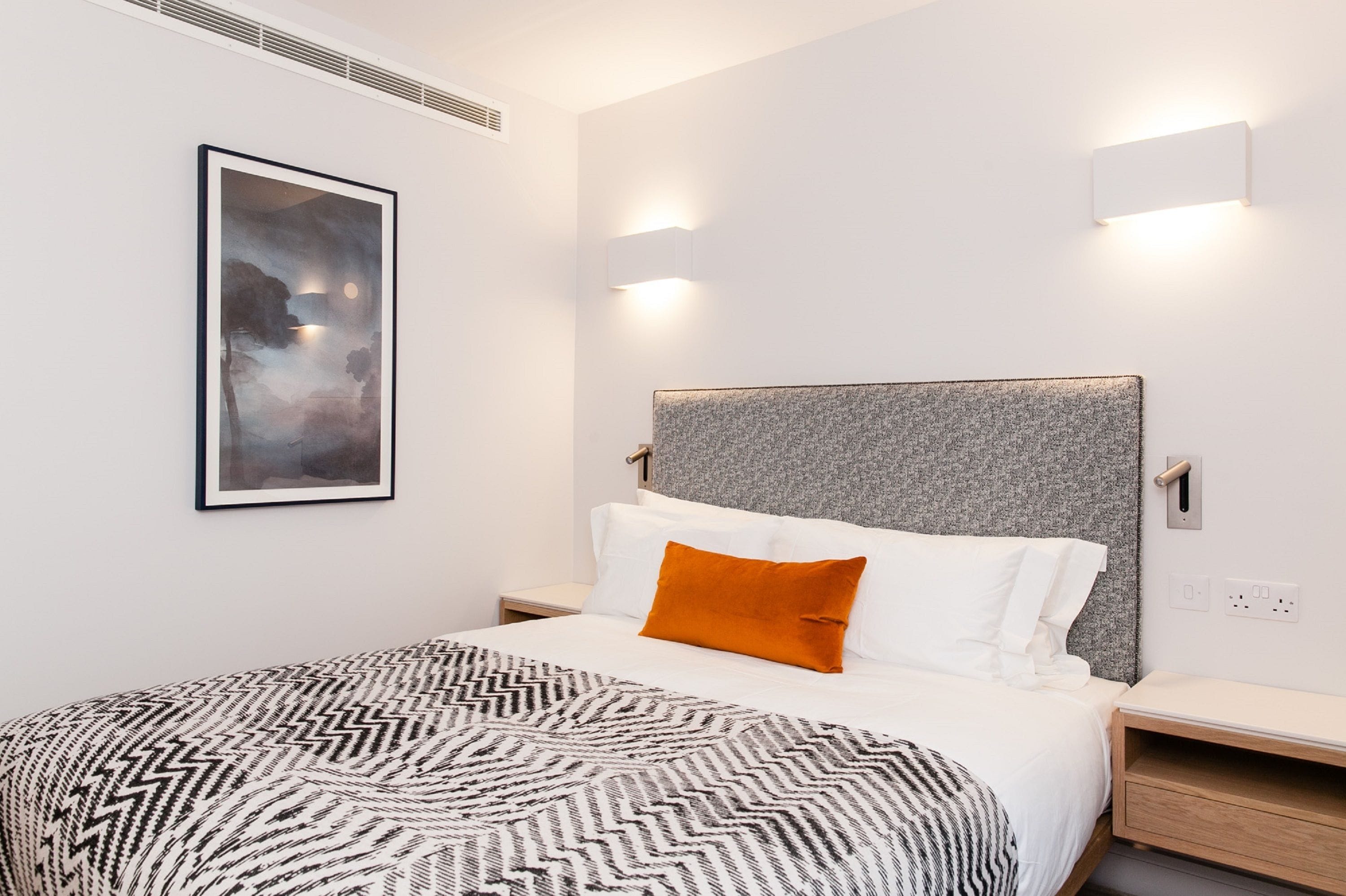 A modern hotel room with a large bed, a headboard with a textured grey panel, a framed abstract painting on the wall, and two bedside lamps.