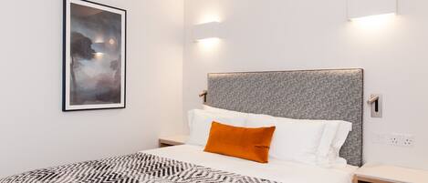 A modern hotel room with a large bed, a headboard with a textured grey panel, a framed abstract painting on the wall, and two bedside lamps.