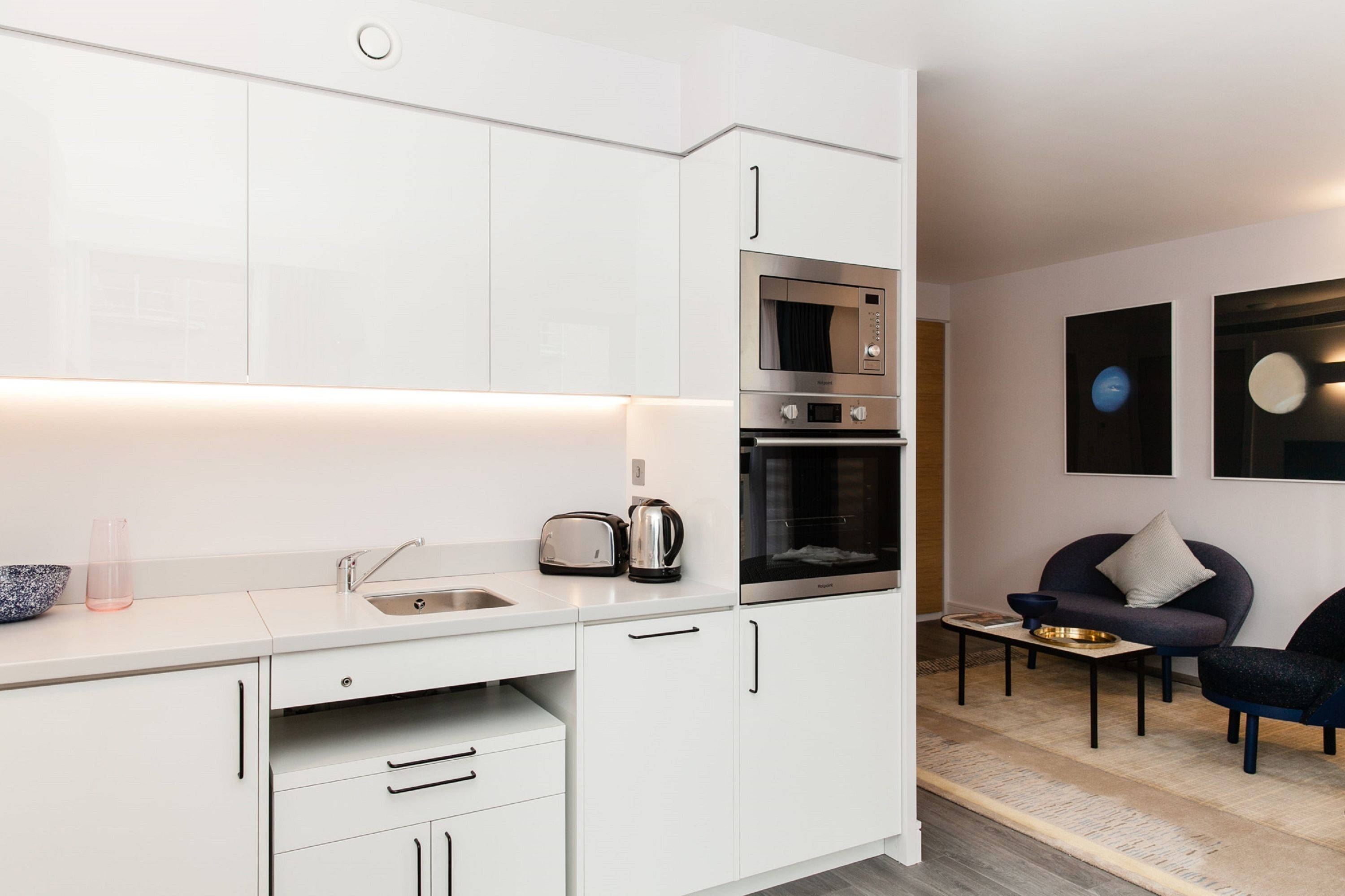 A modern kitchen with white cabinetry, a built-in oven, and a microwave. There is a small dining area with a table and chairs.