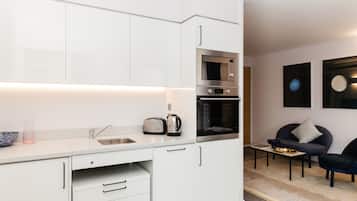 A modern kitchen with white cabinetry, a built-in oven, and a microwave. There is a small dining area with a table and chairs.