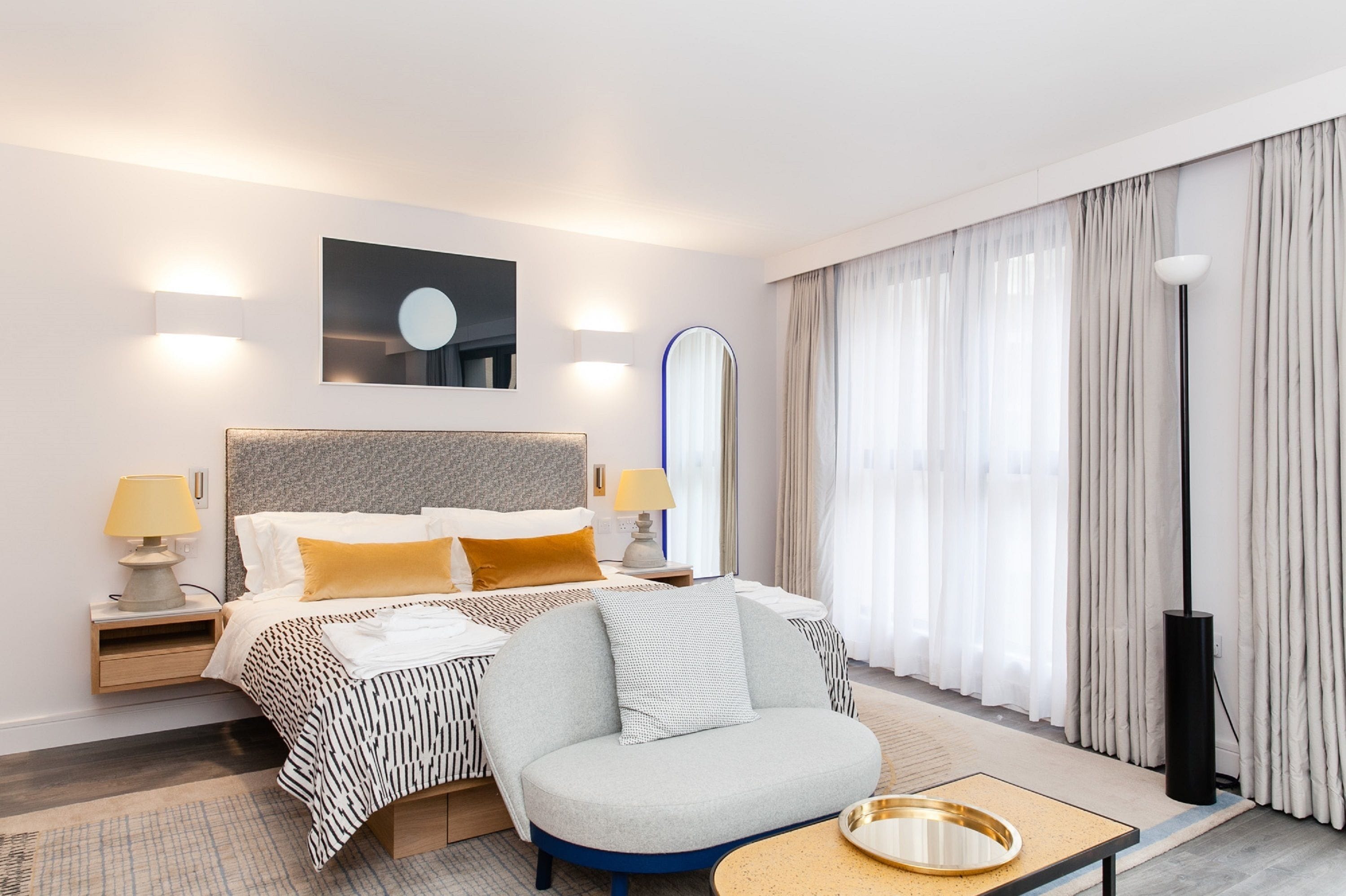 A modern hotel room with a large bed, two bedside tables with lamps, a round mirror, a sofa, and a large window with curtains.