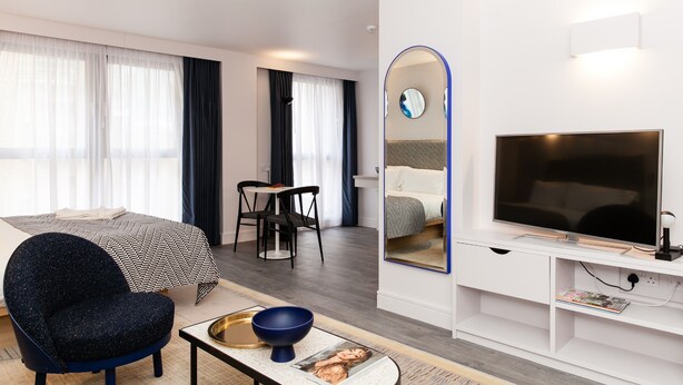 A modern hotel room with a bed, a desk, a chair, a TV, and a mirror.