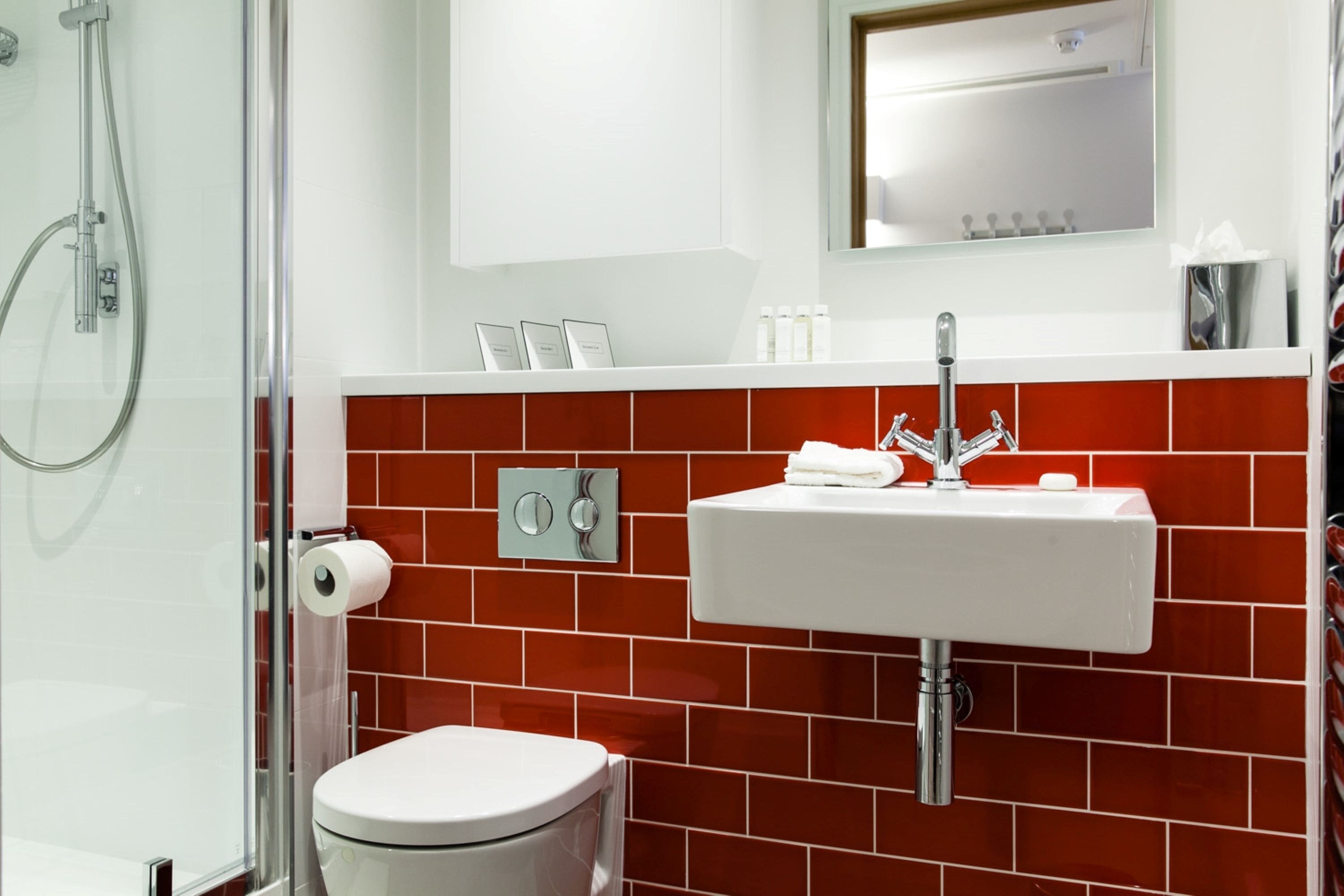 Une salle de bain moderne avec des toilettes blanches, une douche en verre, un mur recouvert de carreaux rouges, un lavabo blanc avec un robinet chromé et un miroir au-dessus du lavabo.