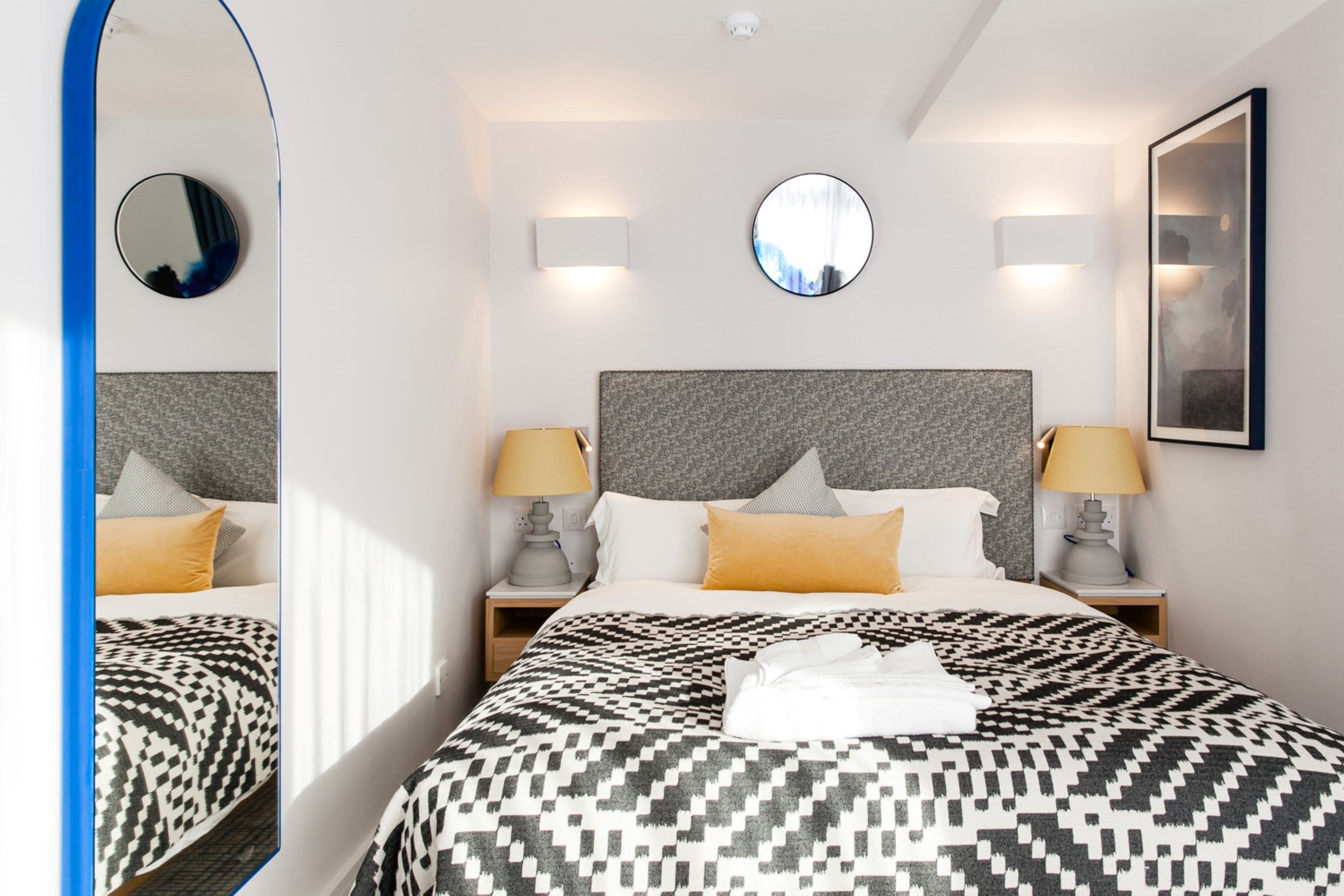 Une chambre d’hôtel avec un grand lit, deux tables de chevet avec des lampes, un miroir rond et un tableau encadré au mur.
