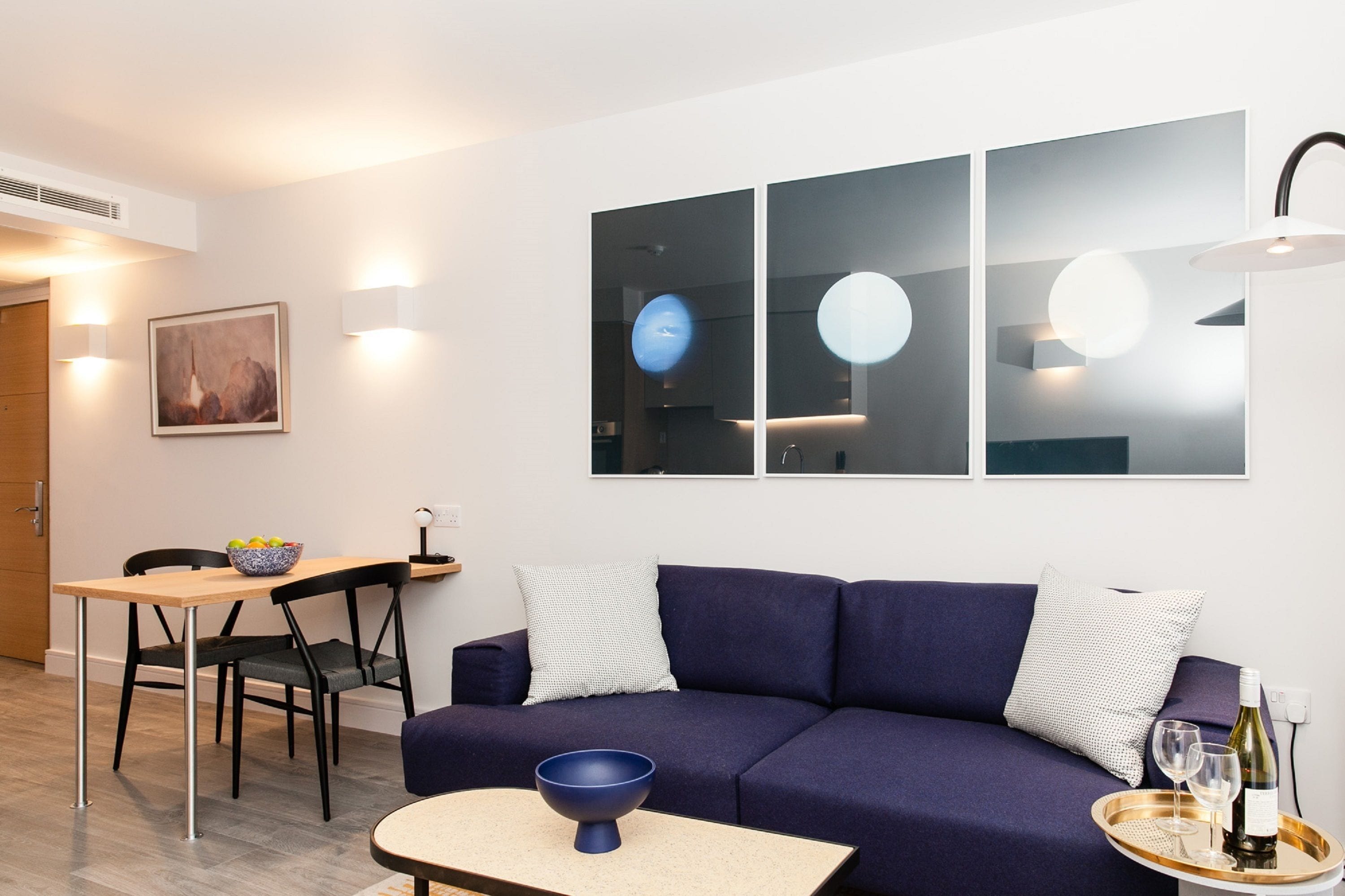A modern living room with a dark blue sofa, a wooden dining table with chairs, and a small coffee table. There are wall-mounted lights and a framed picture on the wall.