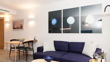 A modern living room with a dark blue sofa, a wooden dining table with chairs, and a small coffee table. There are wall-mounted lights and a framed picture on the wall.