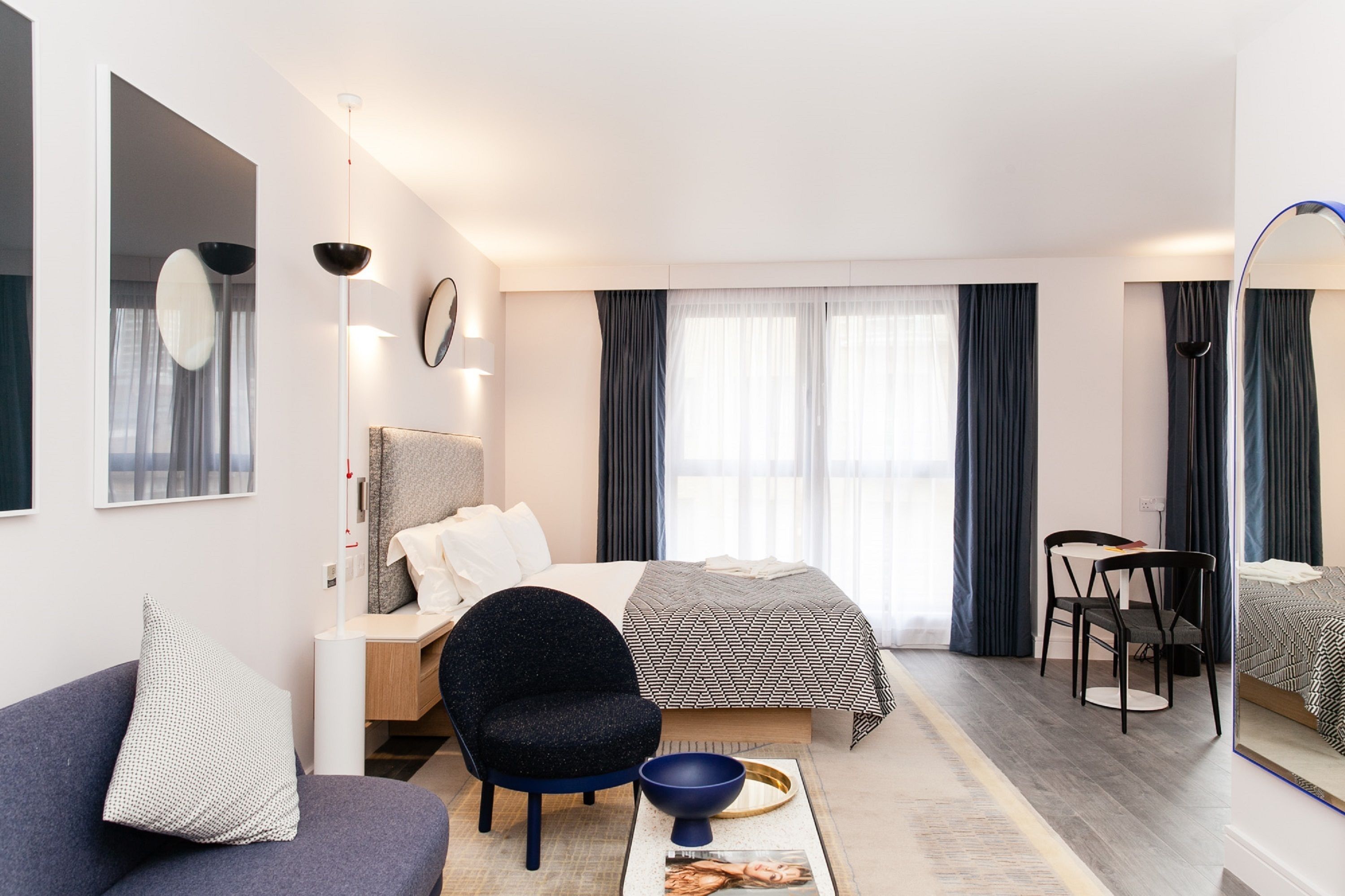 Una moderna camera d'albergo con un letto, una zona salotto con divano e poltrone, un tavolo da pranzo con una ciotola e un grande specchio.
