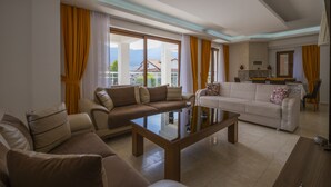 A spacious living room with a large glass coffee table, two sofas, and a dining area with a kitchen in the background. - Infinity Olympia Villas (Fethiye)