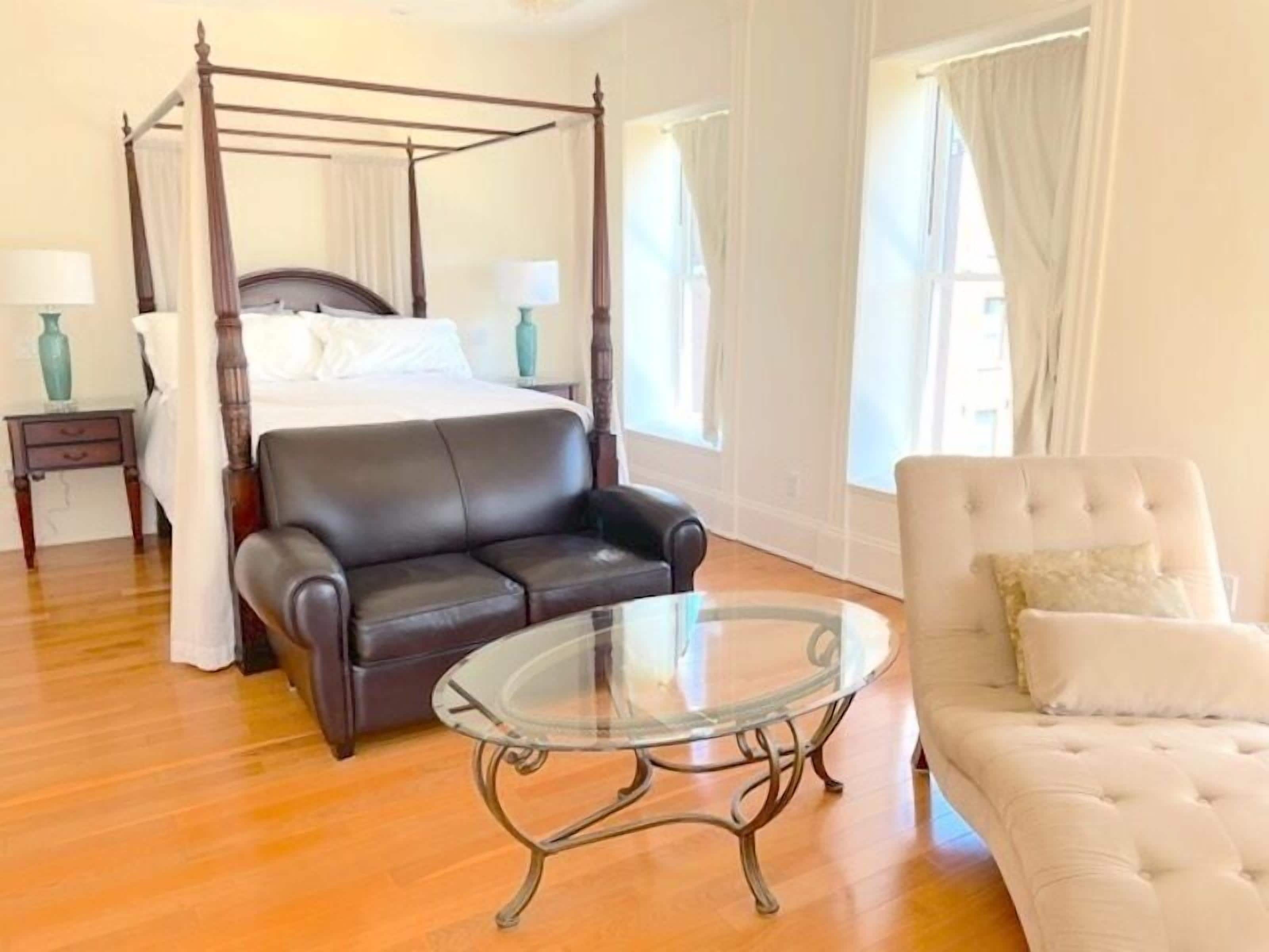 A four-poster bed, a leather sofa, a glass coffee table, and a tufted armchair in a room with wooden flooring and white walls.
