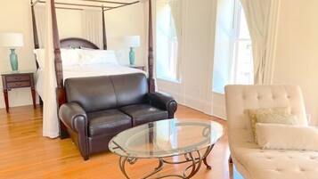 A four-poster bed, a leather sofa, a glass coffee table, and a tufted armchair in a room with wooden flooring and white walls.