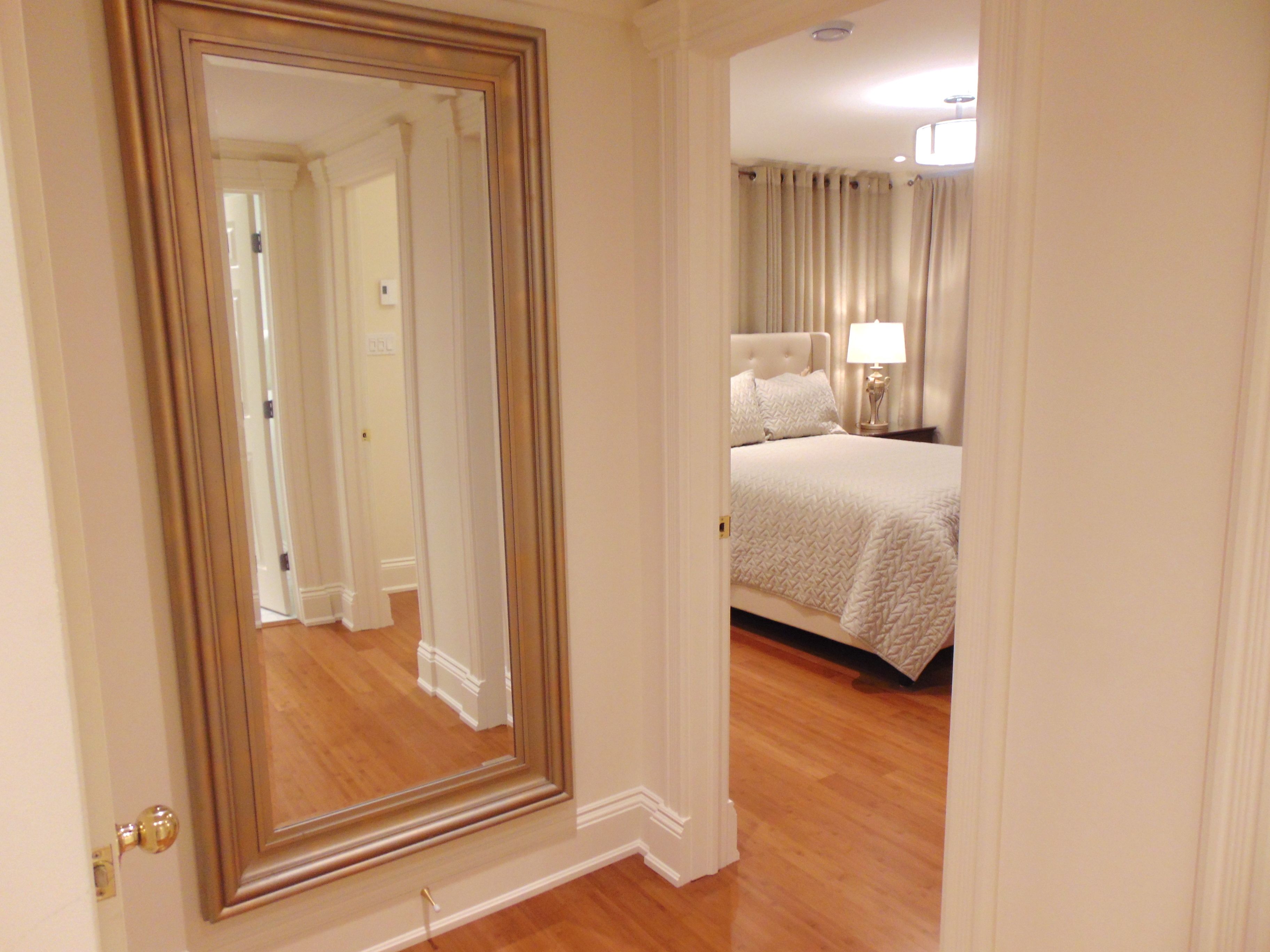 A hotel room with a large mirror, a bed with white linens, a nightstand with a lamp, and wooden flooring.