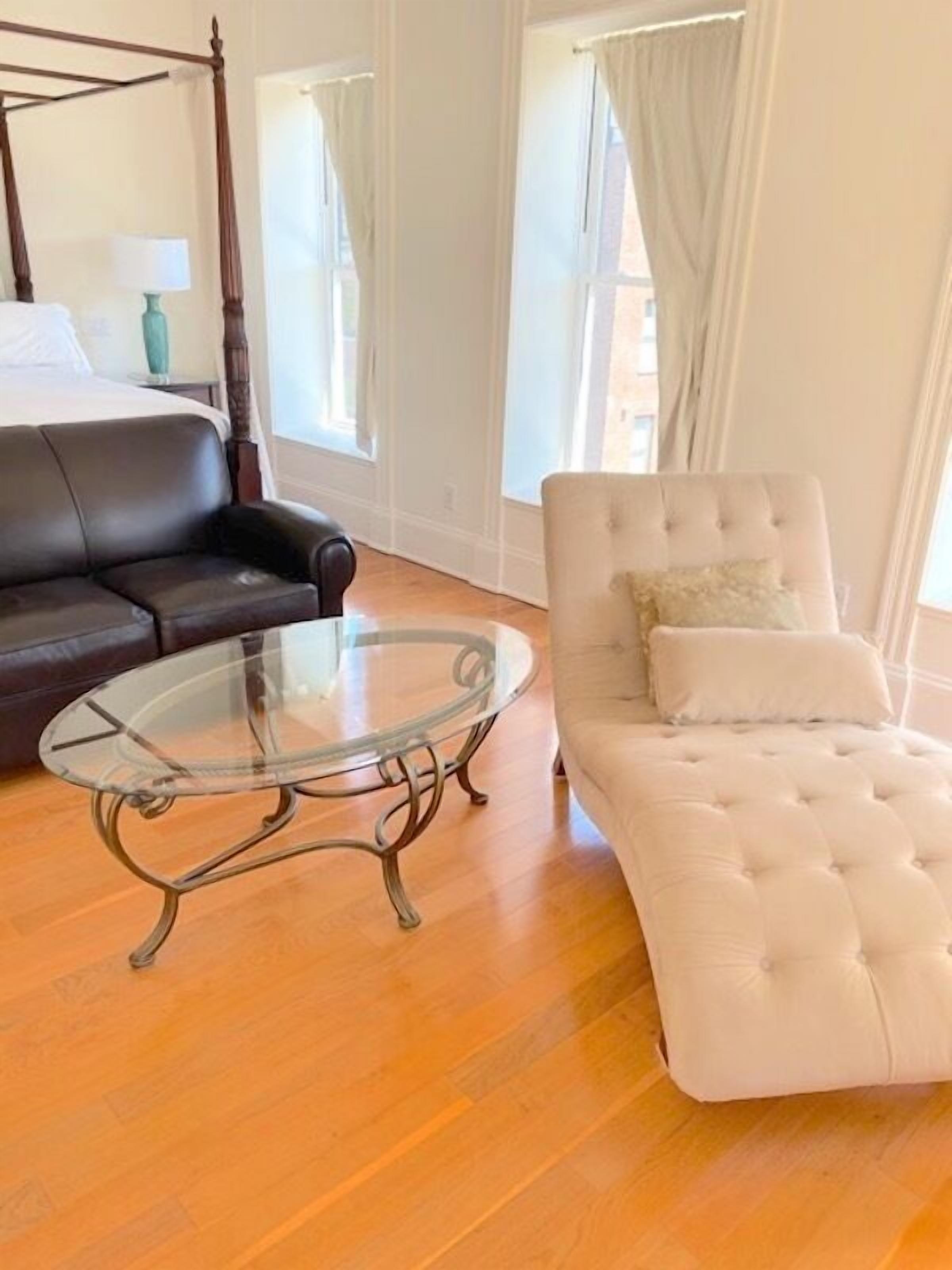 A bedroom with a four-poster bed, a dark leather sofa, a glass coffee table, and a tufted white sofa.